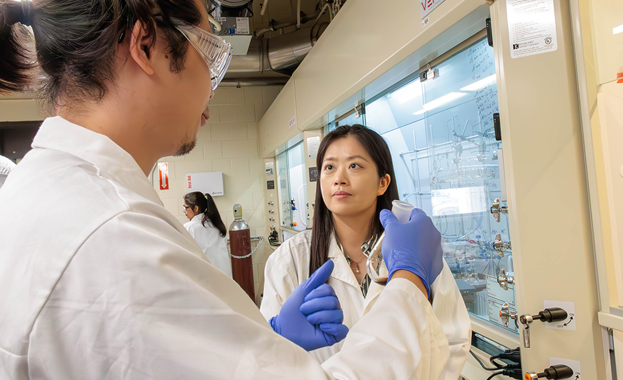 Two scientists in lab coats and safety glasses are discussing work in a chemical laboratory filled with glassware and equipment.