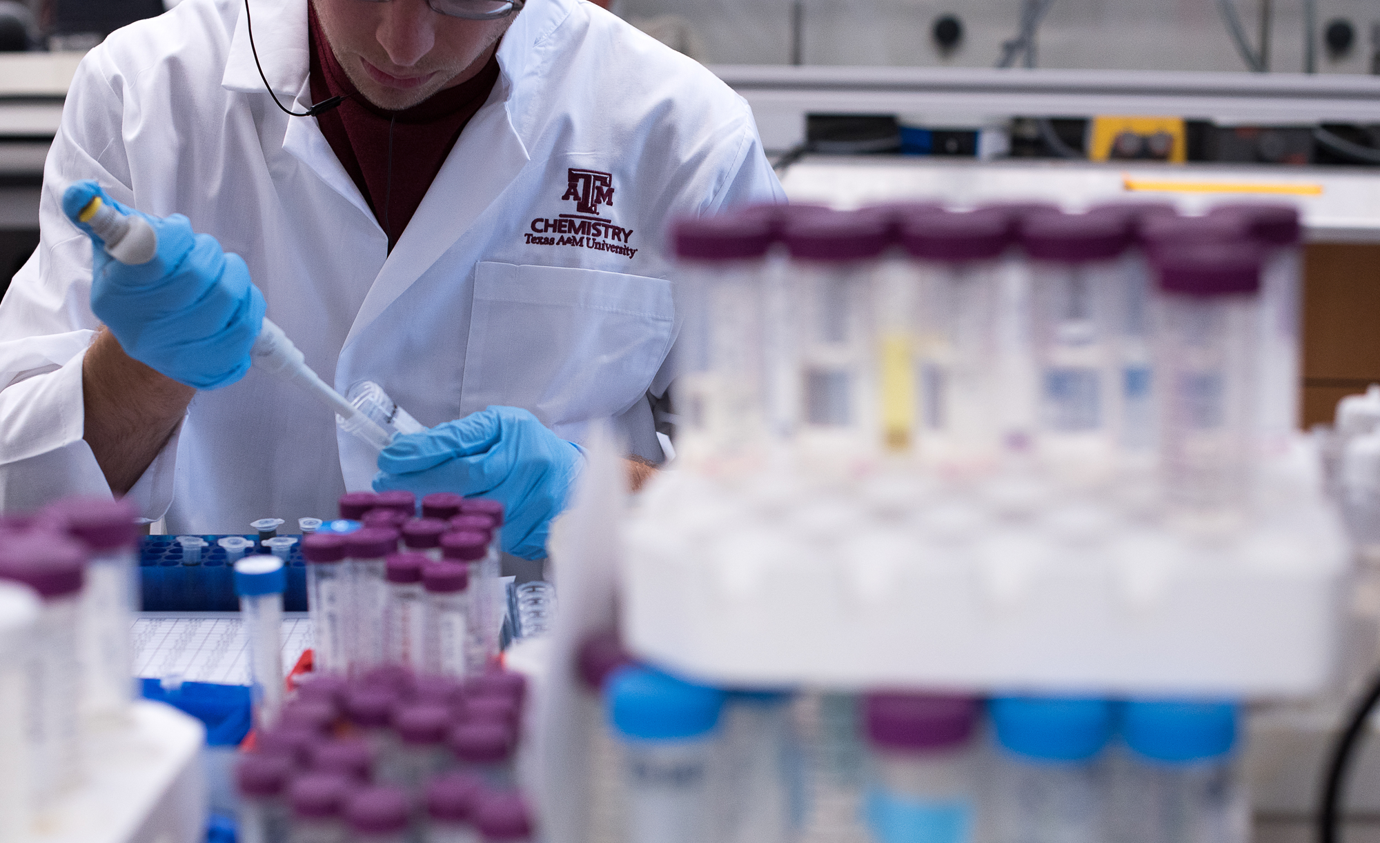 A Texas A&amp;M University lab technician in a white lab coat labeled "Chemistry" is pipetting a substance into a test tube, surrounded by racks of samples.