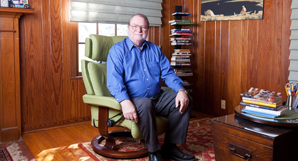 An individual seated in a green armchair in a wood-paneled room, with a stack of books to one side and a painting above. A toy car is on a shelf in the background.