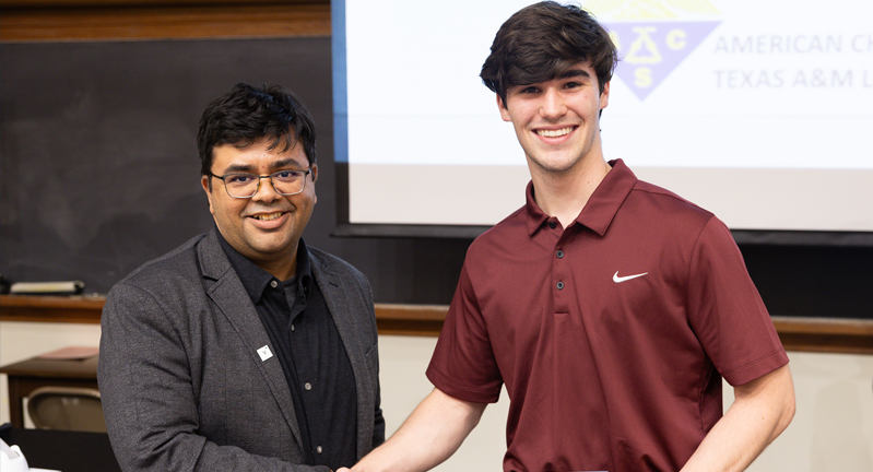 Two people shaking hands in front of a classroom with a banner reading "American Chemical Society Texas A&amp;M University." One person is wearing a dark suit and the other a maroon polo shirt with a Nike logo.