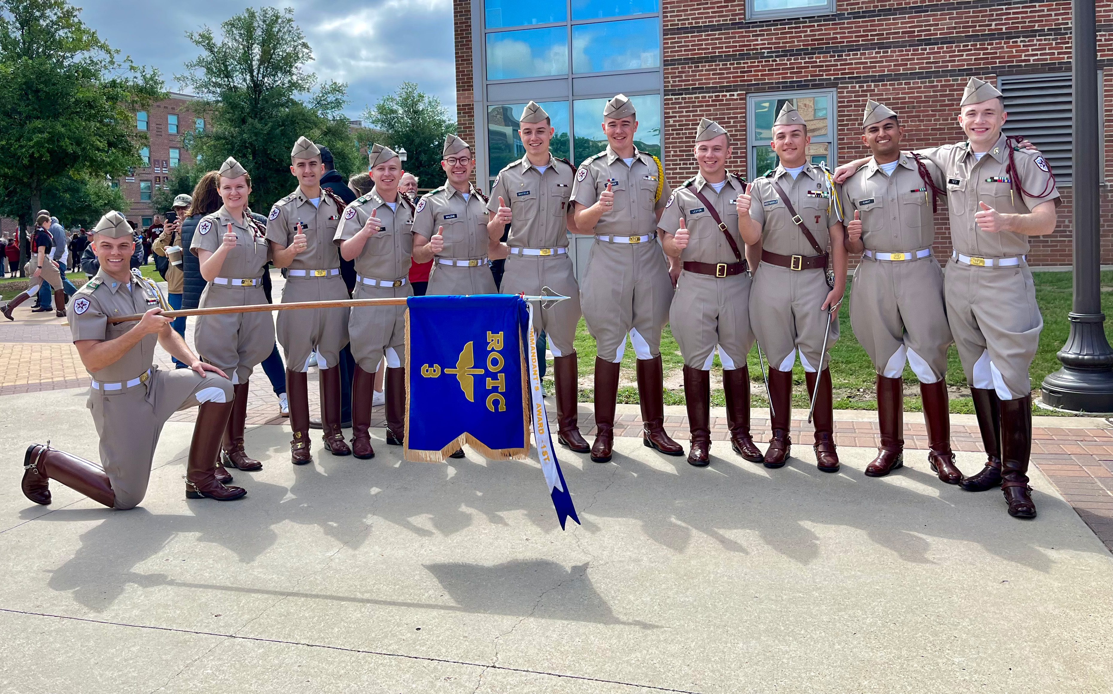 Group of cadets in uniform from Texas A&M University Corps of Cadets standing together outdoors, holding a blue banner with their unit insignia.