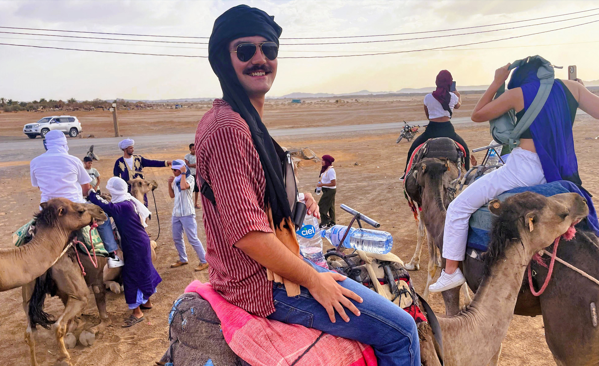 Person smiling while sitting on a camel, with other riders and camels in the background under a clear sky.