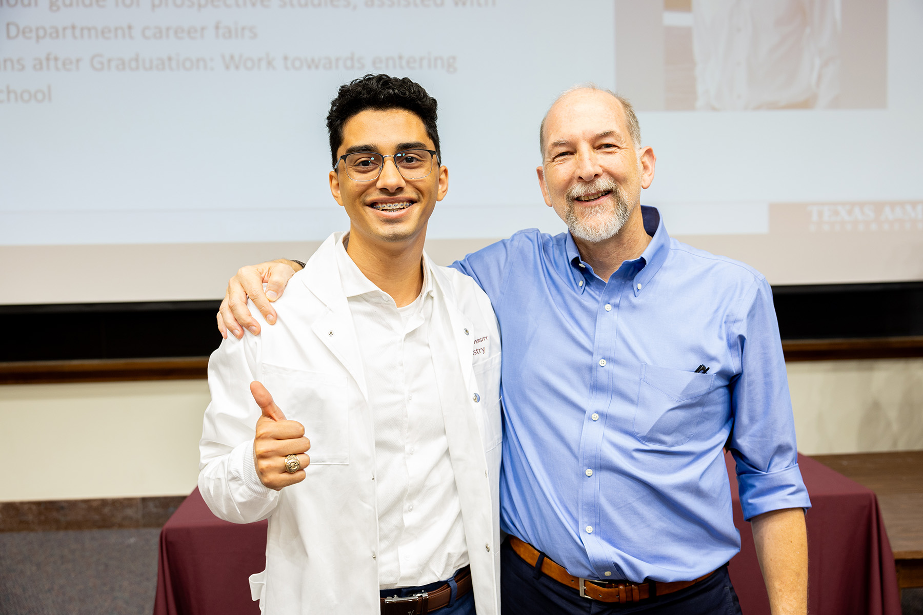 Two people, one in a white coat and giving a thumbs up, smile at the camera.