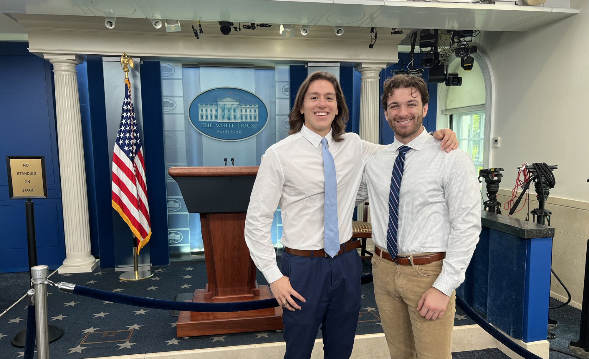 Two individuals smiling in the White House press briefing room, standing next to the speaker's podium with the U.S. flag in the background.
