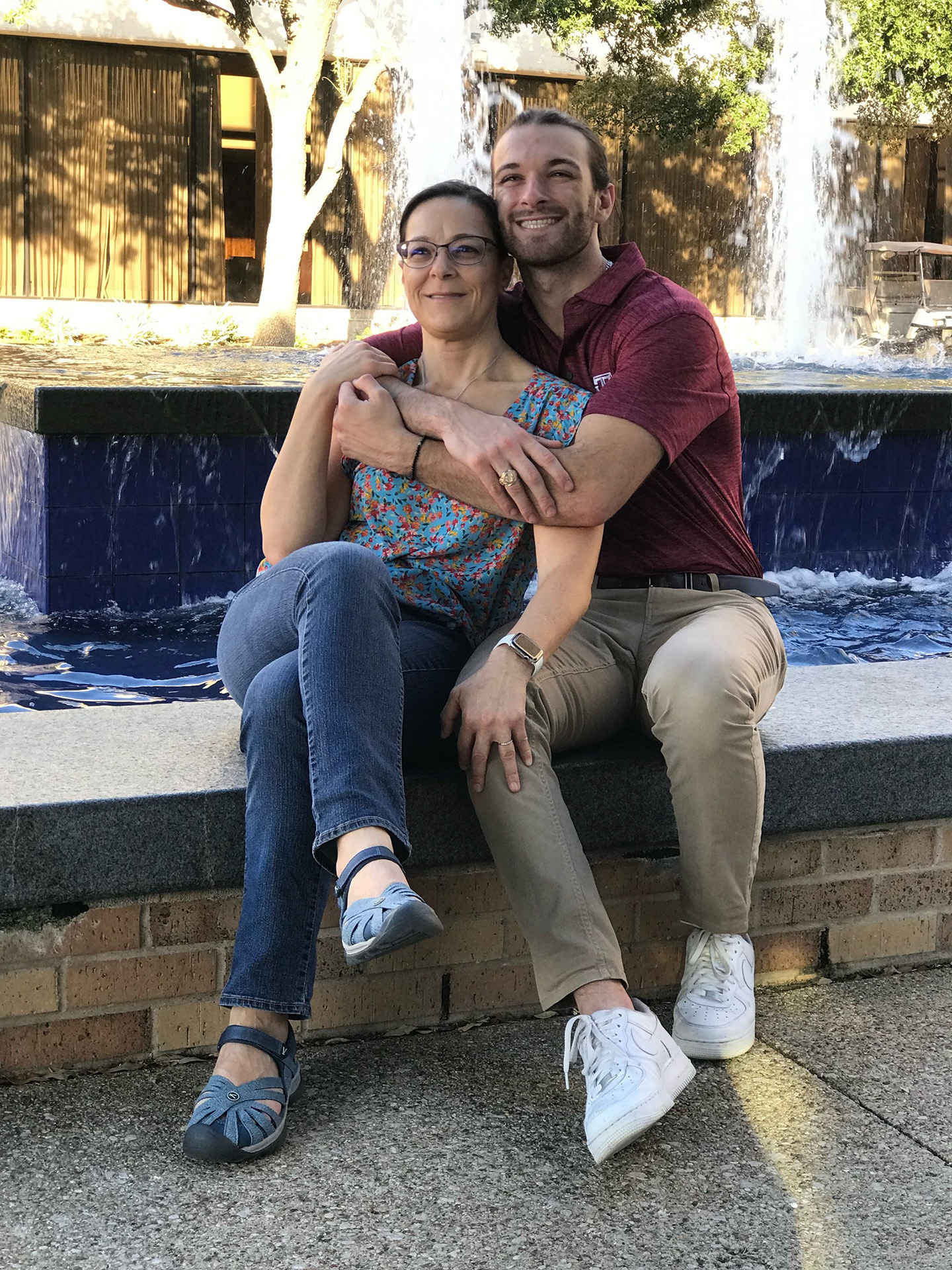 Eric and his mom sit on the ledge of the MSC fountain. Eric hugs his mom from behind, and they are smiling.