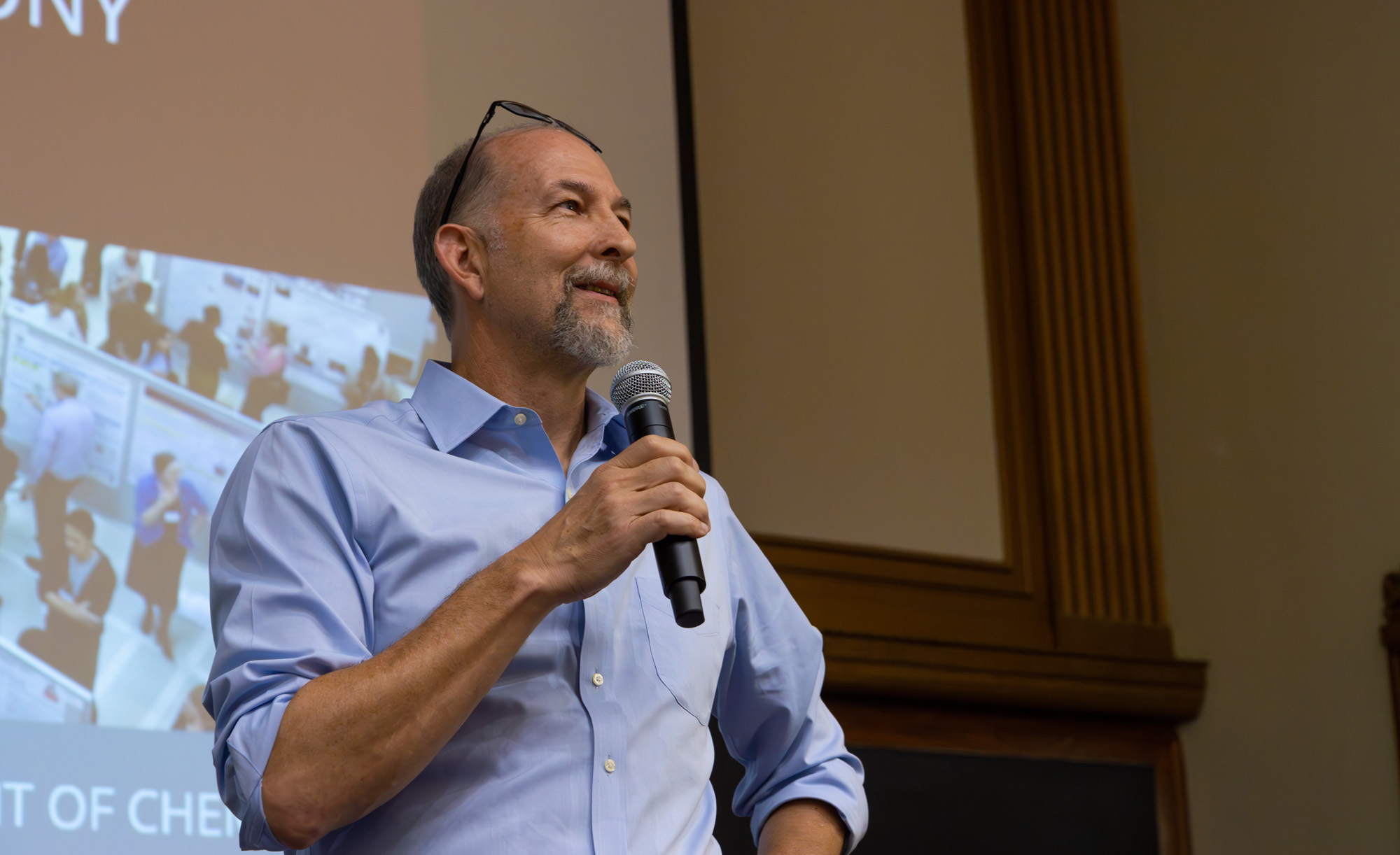 A person speaking at a podium with a microphone in hand, in a room with a projected presentation in the background titled "Department of Chemistry." The individual is wearing a blue shirt and glasses.