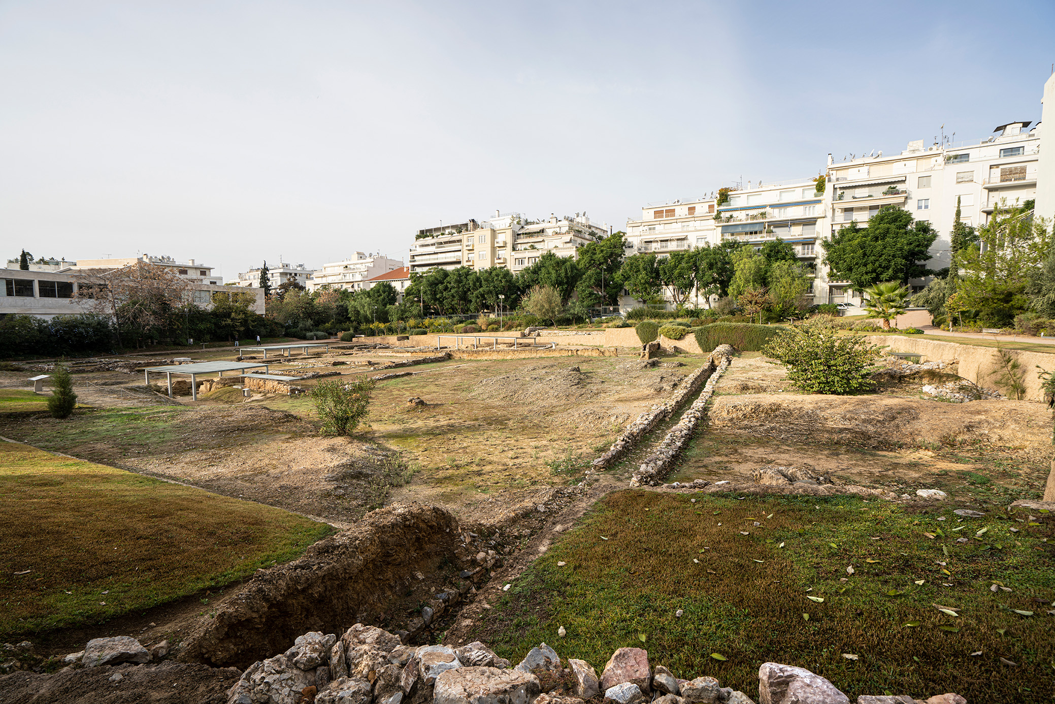 Archaeological site in an urban setting with excavated ruins, surrounded by modern apartment buildings.
