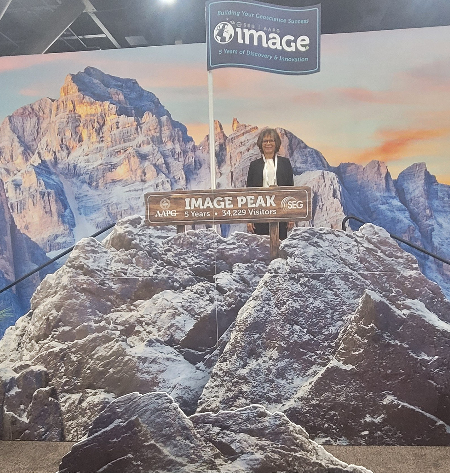 Dr. Sherilyn Williams-Stroud stands behind a sign reading “IMAGE PEAK, AAPG SEG, 5 Years, 34,229 Visitors” at a geoscience conference exhibit. A scenic mountain backdrop and a banner above highlight five years of discovery and innovation in geoscience.