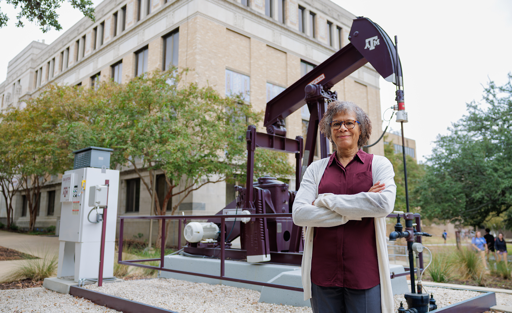 A person stands with arms crossed in front of a large oil pump jack bearing the Texas A&M University logo, set on a university campus with a beige brick building and surrounding greenery in the background