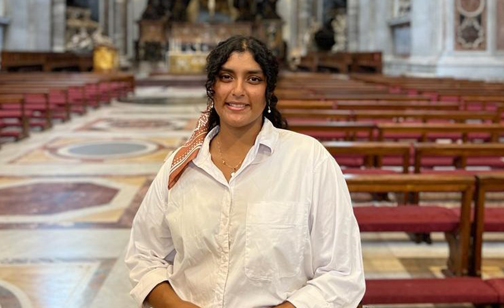Person wearing a white button-down shirt and a patterned scarf tied around the hair, standing in the apse of St. Peter’s Basilica in Vatican City. The setting includes marble floors with geometric designs, rows of wooden benches with red cushions, tall columns, and ornate architectural details. In the background, there is an altar area featuring gold accents and intricate decorations.