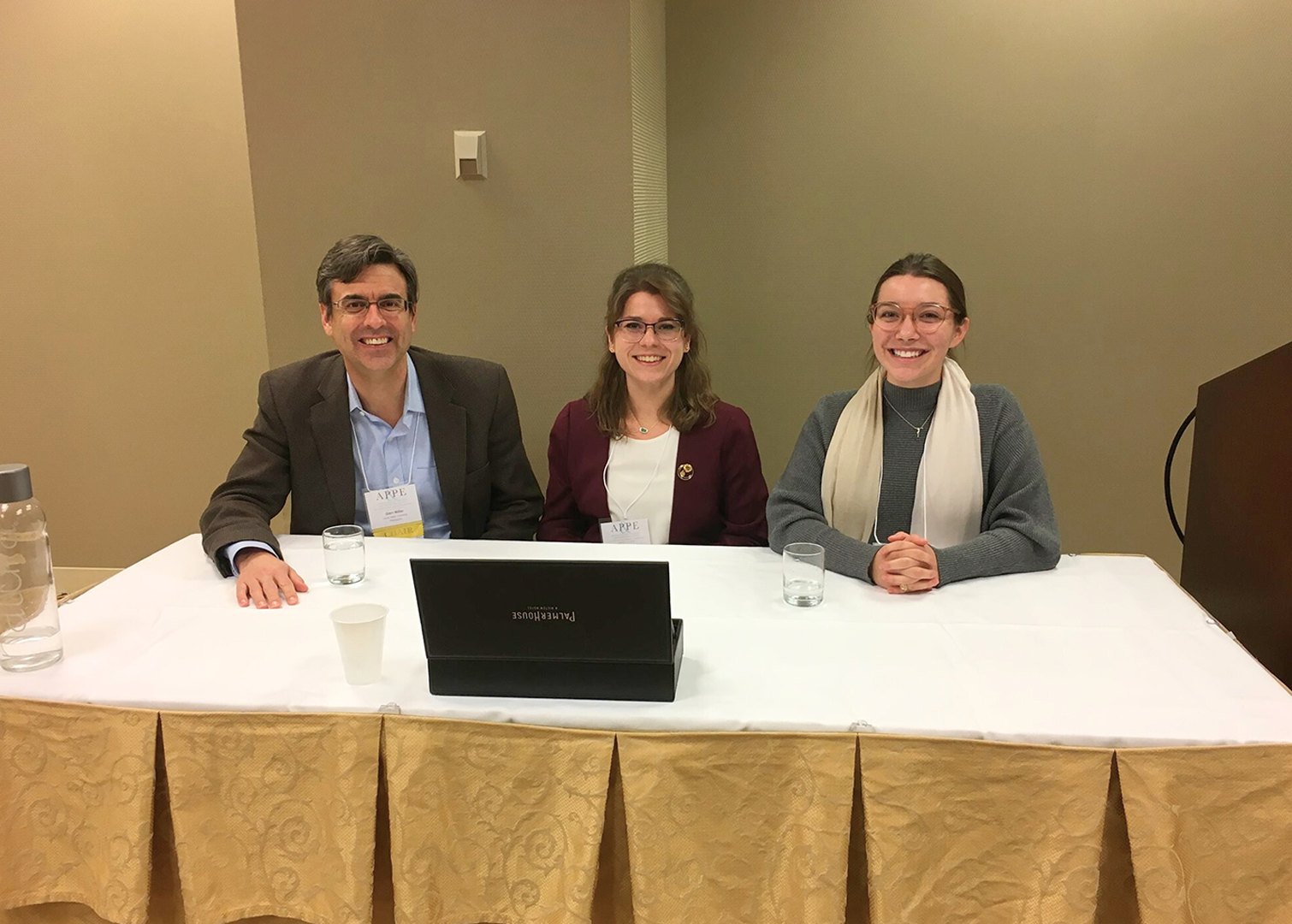 Three people seated at a table with a white tablecloth and gold skirt, with a laptop, water pitcher, and cups on the table. A podium is partially visible to the right against a beige wall.