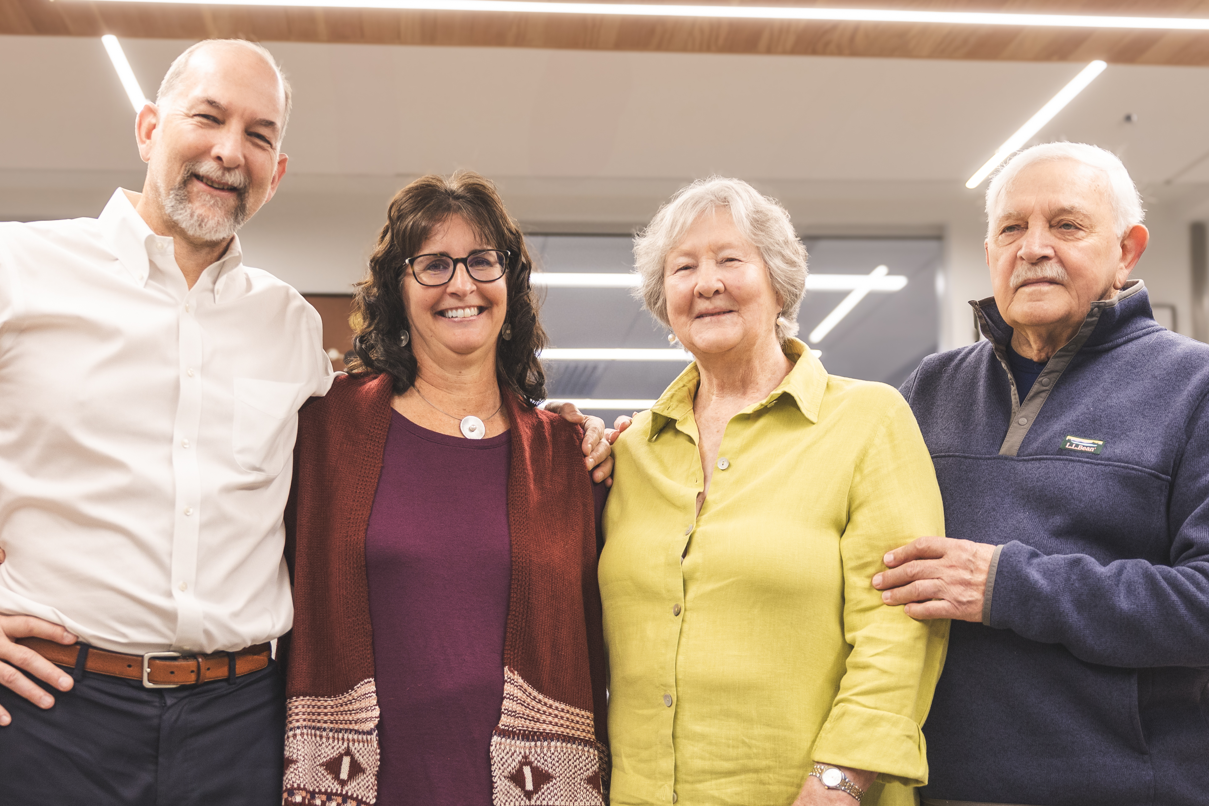 Four people standing side by side indoors, with arms around each other. They are dressed in casual to semi-formal clothing: one in a white button-down shirt, another in a maroon top with a patterned cardigan, one in a light green shirt, and another in a dark fleece pullover. The background shows a modern interior with bright overhead lights and wooden ceiling panels