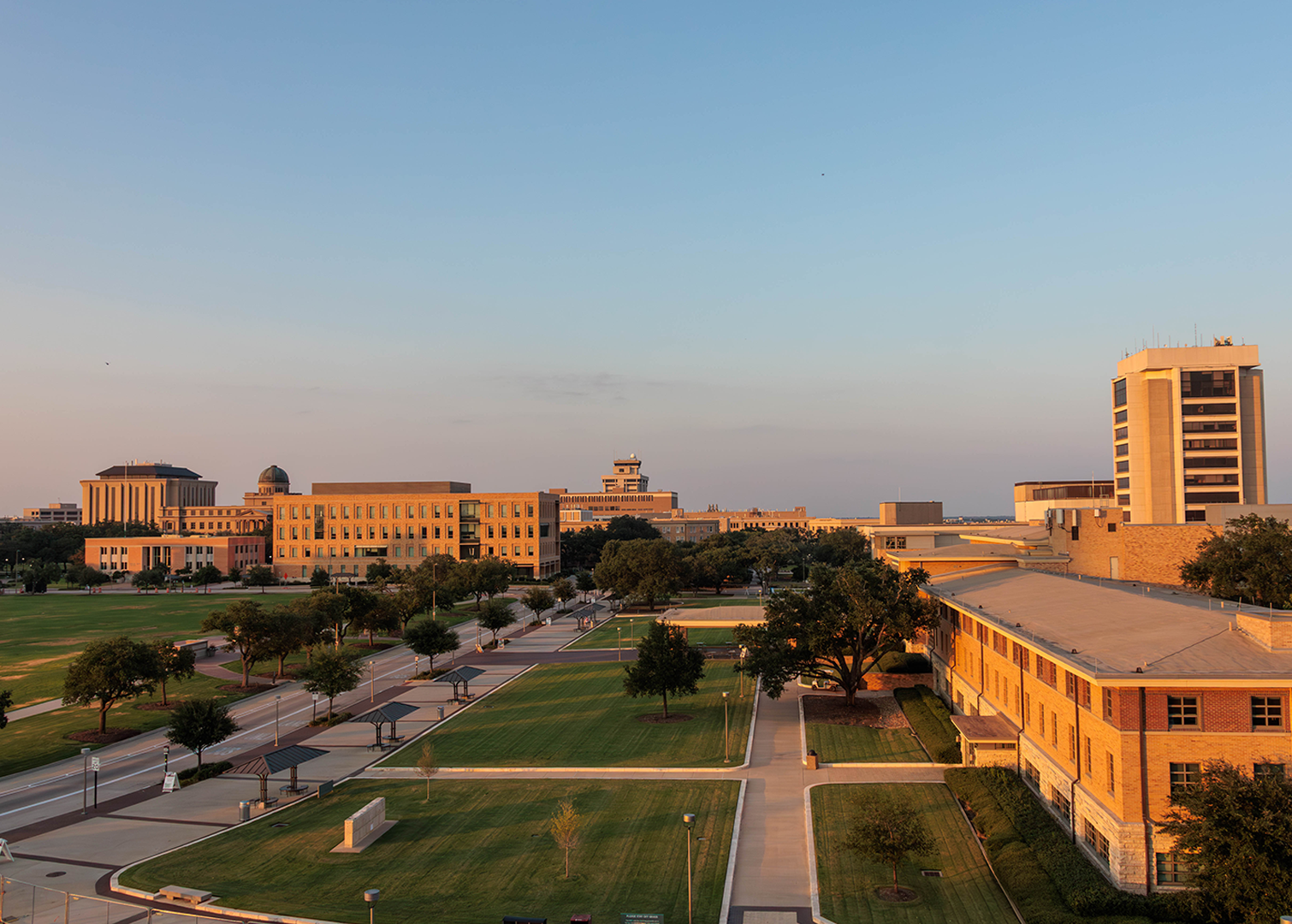 Wide view of Texas A&amp;M University campus buildings at sunset, featuring tree-lined walkways, green lawns, and academic buildings with tan brick exteriors under a clear blue sky.