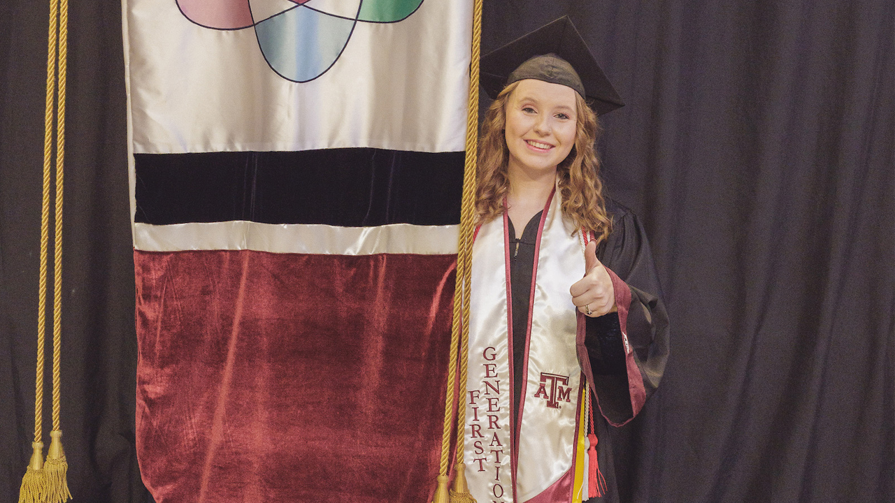 Megan in a black graduation gown and cap smiling proudly, wearing a "First Generation" stole and honor cords at a Texas A&M University graduation ceremony, with the gonfalon to her left.