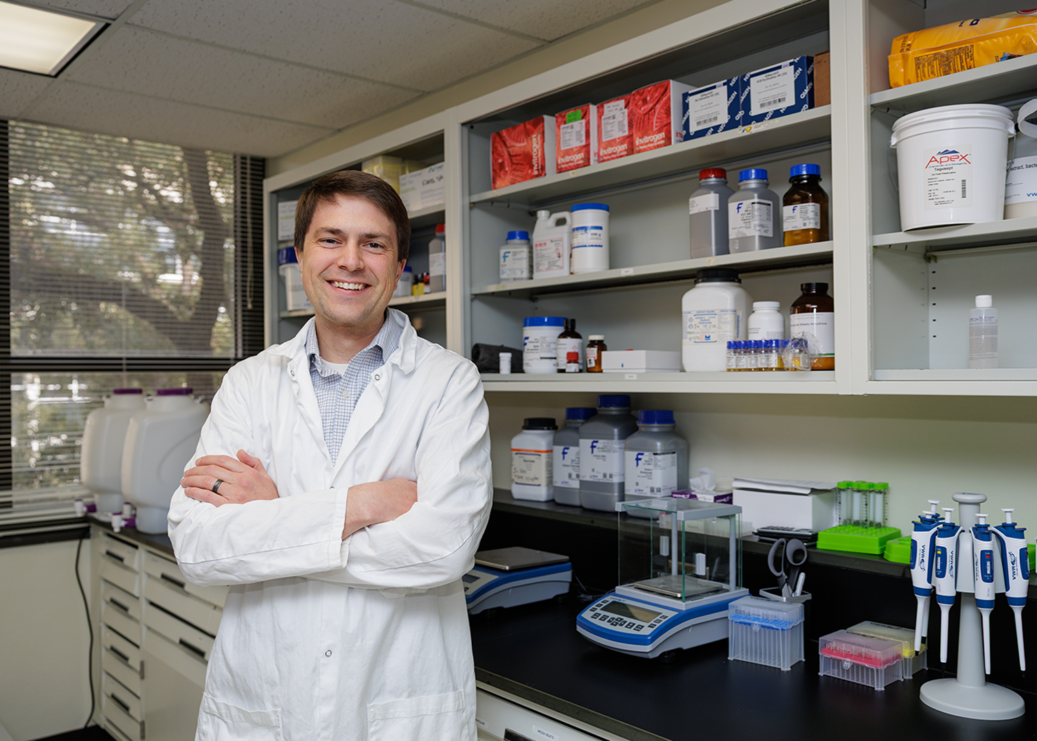 Person wearing a white lab coat standing with arms crossed in a laboratory. Behind them are shelves stocked with chemical containers, lab supplies, and equipment. The countertop holds pipettes, scales, and other scientific instruments, with a window showing trees in the background.