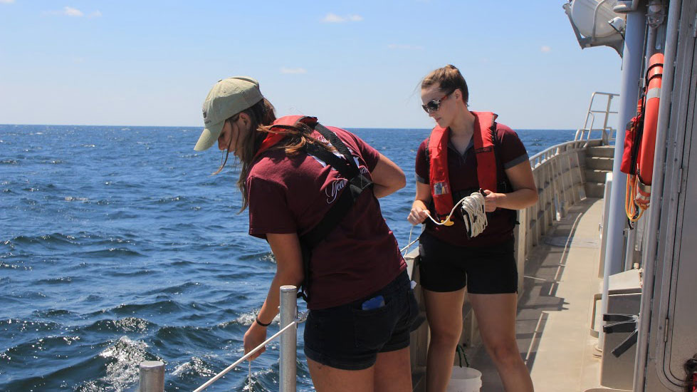 Two students guide a rope over the side of a boat out at sea.