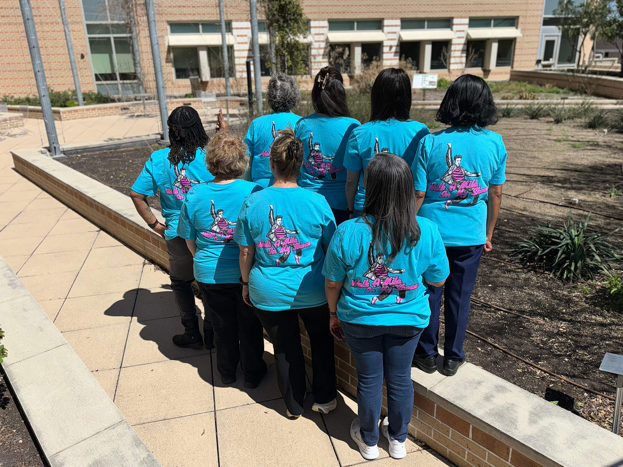 Eight women standing near a rooftop garden with their backs turned towards the camera, all wearing shirts that read "Walk A Mile In My Shoes" and Elvis Presley.