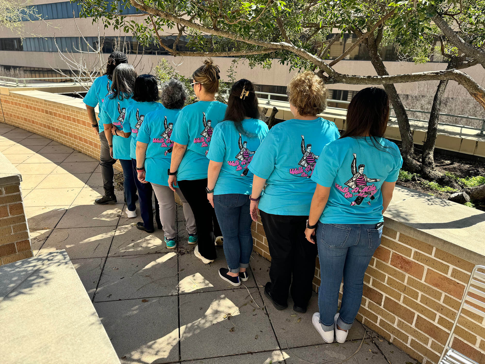 Eight women standing with their backs turned towards the camera, all wearing blue shirts that read "Walk A Mile In My Shoes" with Elvis Presley striking a pose.