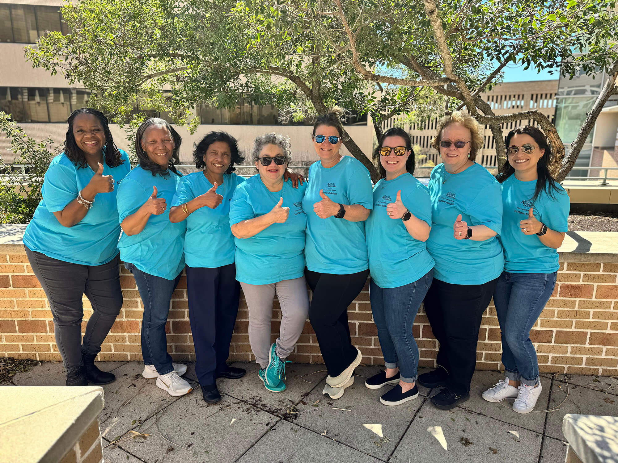 Eight women standing and giving a thumbs up towards the camera, all wearing blue shirts for their team.