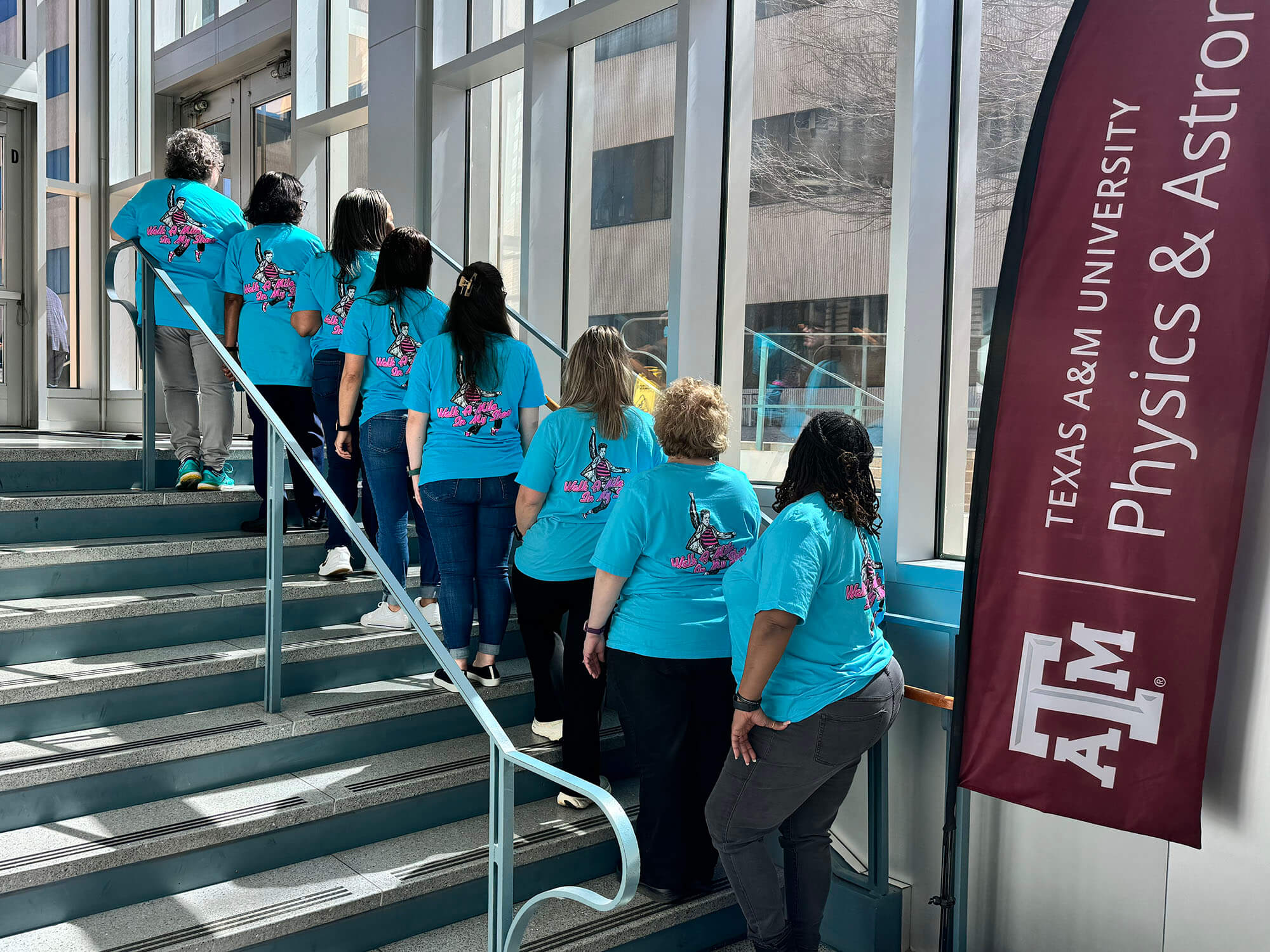 Eight women standing on a stairway with their backs turned towards the camera, all wearing shirts that read "Walk A Mile In My Shoes" and Elvis Presley.