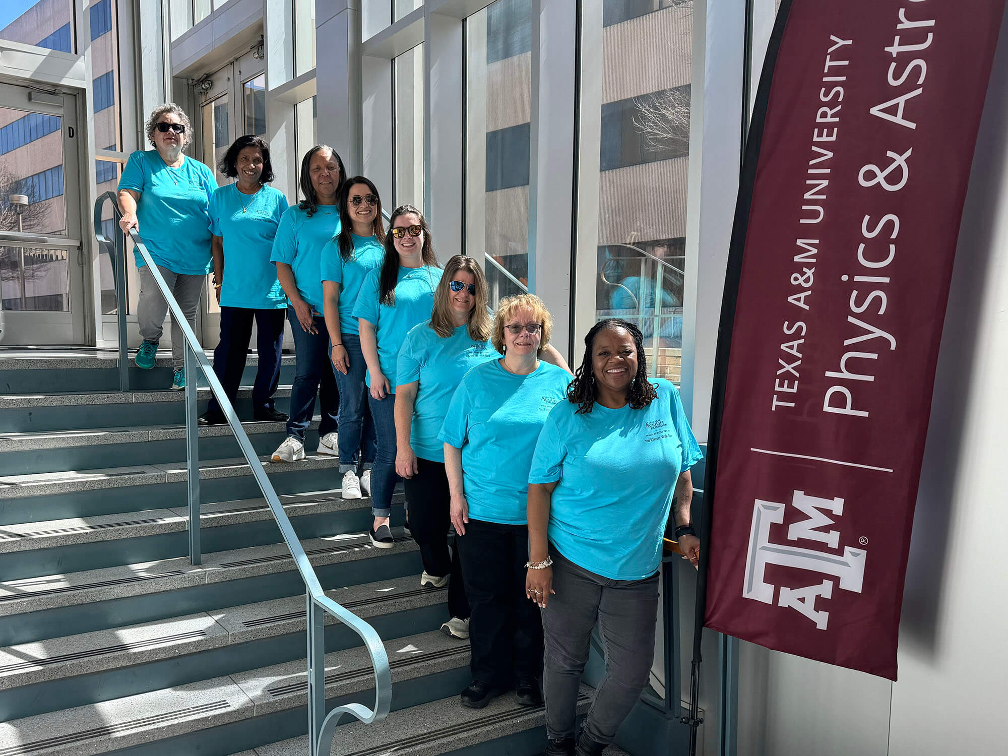 Eight women standing on a stairway and smiling, all wearing blue shirts to represent their team.