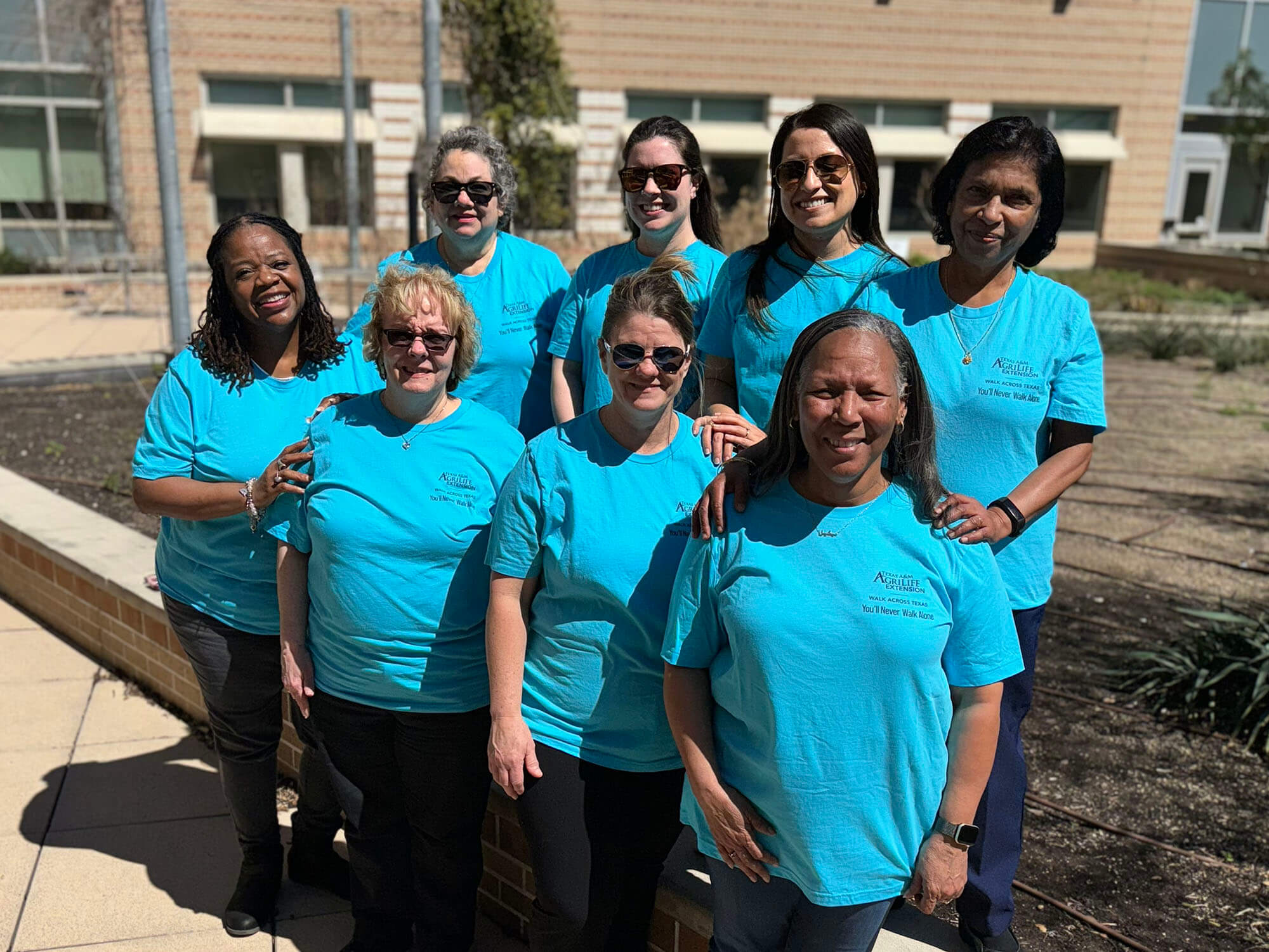 Eight women standing and smiling near a rooftop garden, all wearing blue shirts to represent their team.