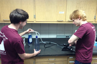 two men at a lab counter. One is holding a flexable rubber tube that is connecting an instrument to a plastic bottle.