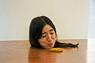 A woman's head stuck through a whole in a wood table top. A cookie is just out of reah, she is pretending to make a sad face because she can't reach the cookie.