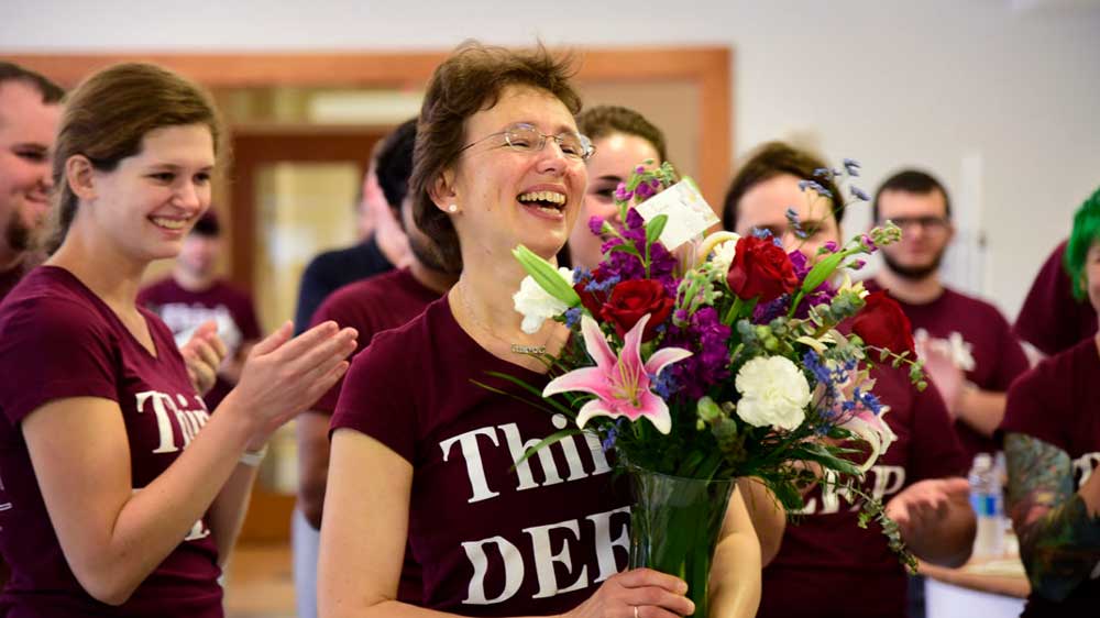 A professor accepts a colorful bouquet of flowers with a big smile while people applaud around her