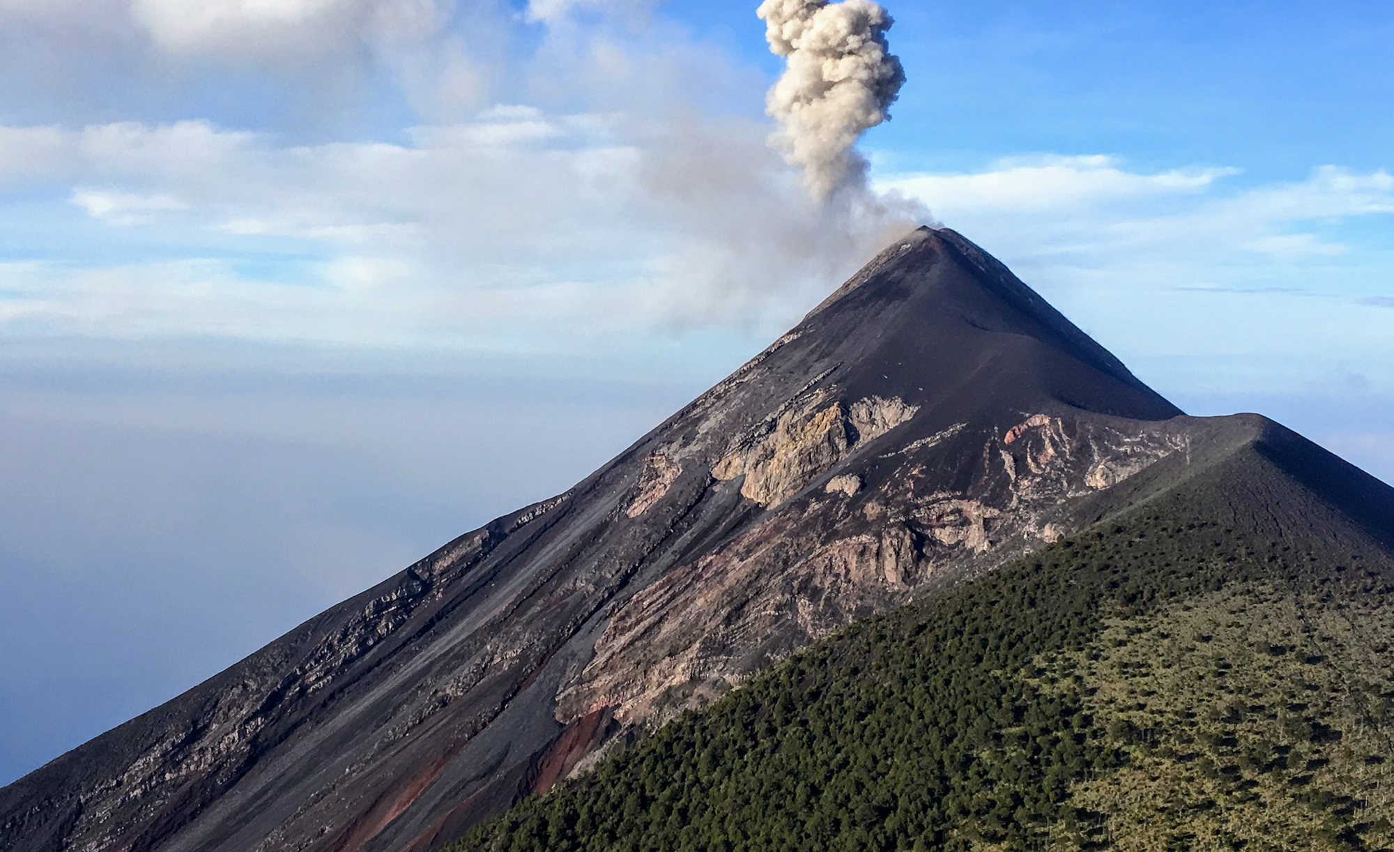 A volcano emitting a plume of smoke against a clear sky, with lush greenery at the base.