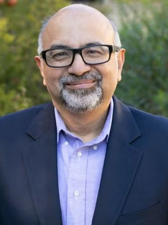 Portrait of Dr. Sudipto Banerjee with glasses, smiling, wearing a blue blazer and a light blue shirt, set against a blurred green foliage background.