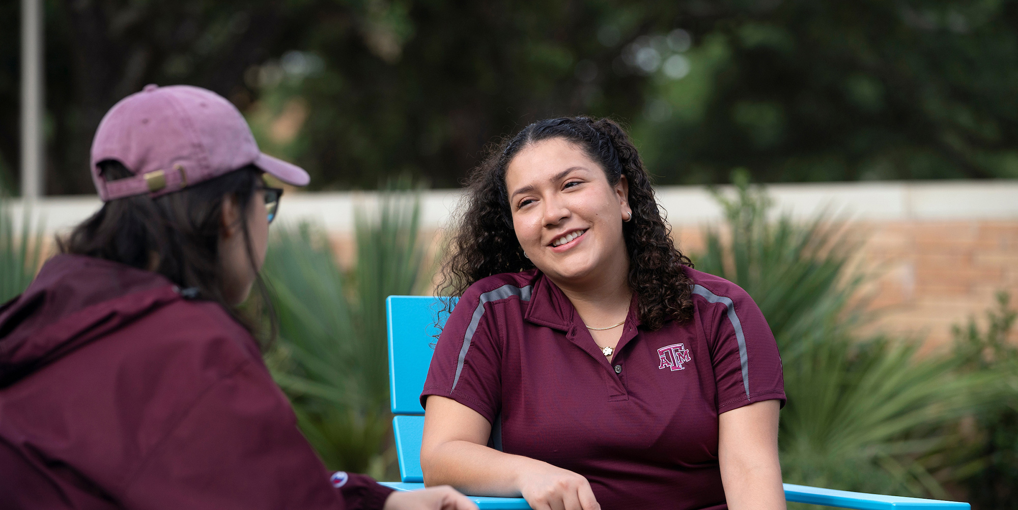 Female student with black hair and maroon Texas A&M polo shirt smiles at another student