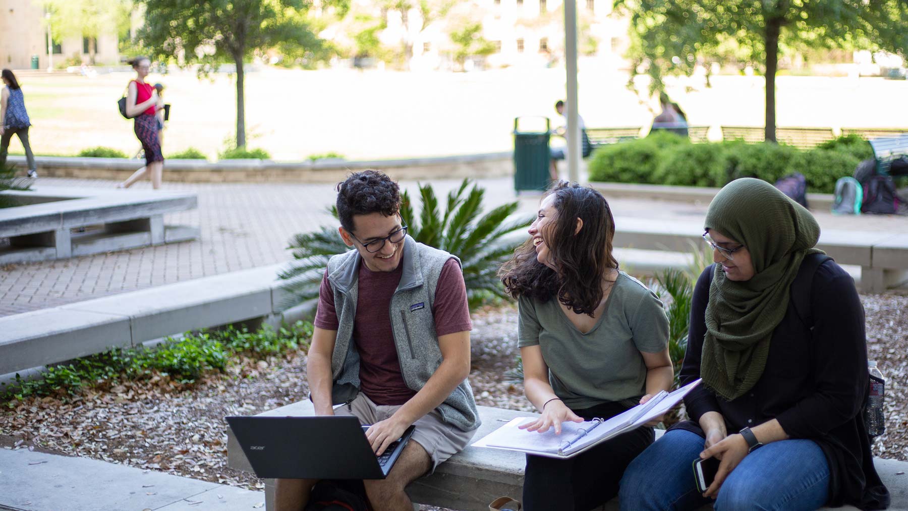 Three Texas A&amp;M students sitting outside the Eller O&amp;M Building, studying together and laughing.